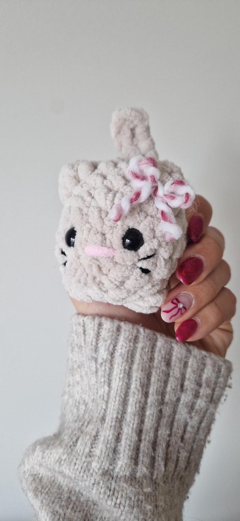 May include: A handmade, light gray crocheted cat-shaped item with black eyes, whiskers, and a pink nose. It has a small bow with red and white stripes. The item is held in a hand.