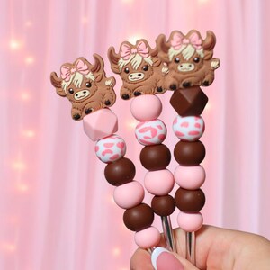 May include: Three decorative skewers with brown Highland cow toppers wearing pink bows. The skewers have pink, brown, and white patterned beads. The background is a soft pink color.