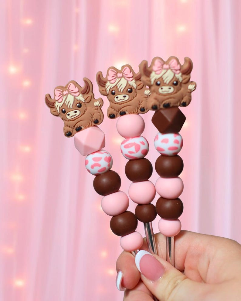 May include: Three decorative skewers with brown Highland cow toppers wearing pink bows. The skewers have pink, brown, and white patterned beads. The background is a soft pink color.