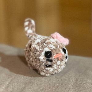 May include: A handmade crochet bunny toy in shades of brown and white, with black eyes and a pink bow. The plush toy has a soft, textured appearance, perfect for children or as a decorative item. The bunny's nose is a light orange color.