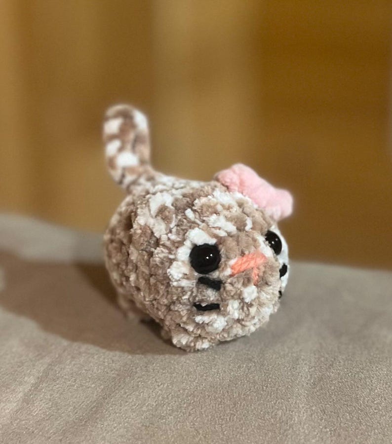 May include: A handmade crochet bunny toy in shades of brown and white, with black eyes and a pink bow. The plush toy has a soft, textured appearance, perfect for children or as a decorative item. The bunny's nose is a light orange color.