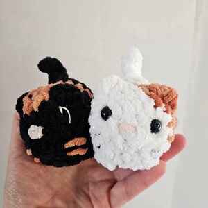 May include: Two crocheted cat-shaped ornaments. One is black with orange and white accents, the other is white with orange and white accents. Both have black bead eyes and small tails. The ornaments are handmade.
