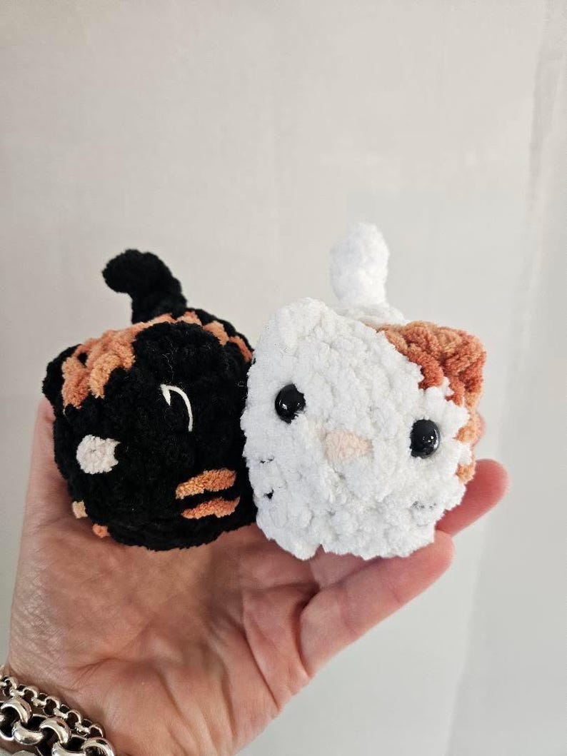 May include: Two crocheted cat-shaped ornaments. One is black with orange and white accents, the other is white with orange and white accents. Both have black bead eyes and small tails. The ornaments are handmade.