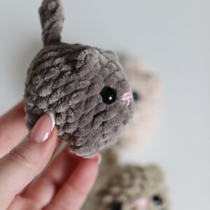 May include: Close-up of three handmade crochet cat toys in shades of gray, beige, and brown. The gray cat is in focus, with black eyes and a pink nose. The toys have a soft, textured appearance, and a small tail.