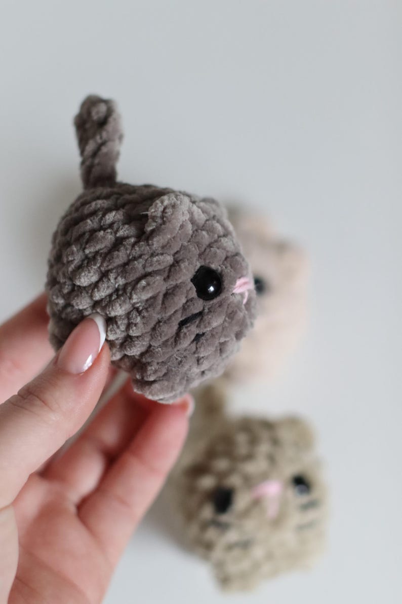 May include: Close-up of three handmade crochet cat toys in shades of gray, beige, and brown. The gray cat is in focus, with black eyes and a pink nose. The toys have a soft, textured appearance, and a small tail.
