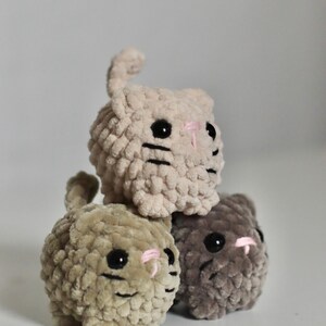 May include: Three handmade, plush cat toys in a stack. The cats are beige, olive green, and gray, with black eyes, pink noses, and black whiskers. The toys are made of a soft, textured material.