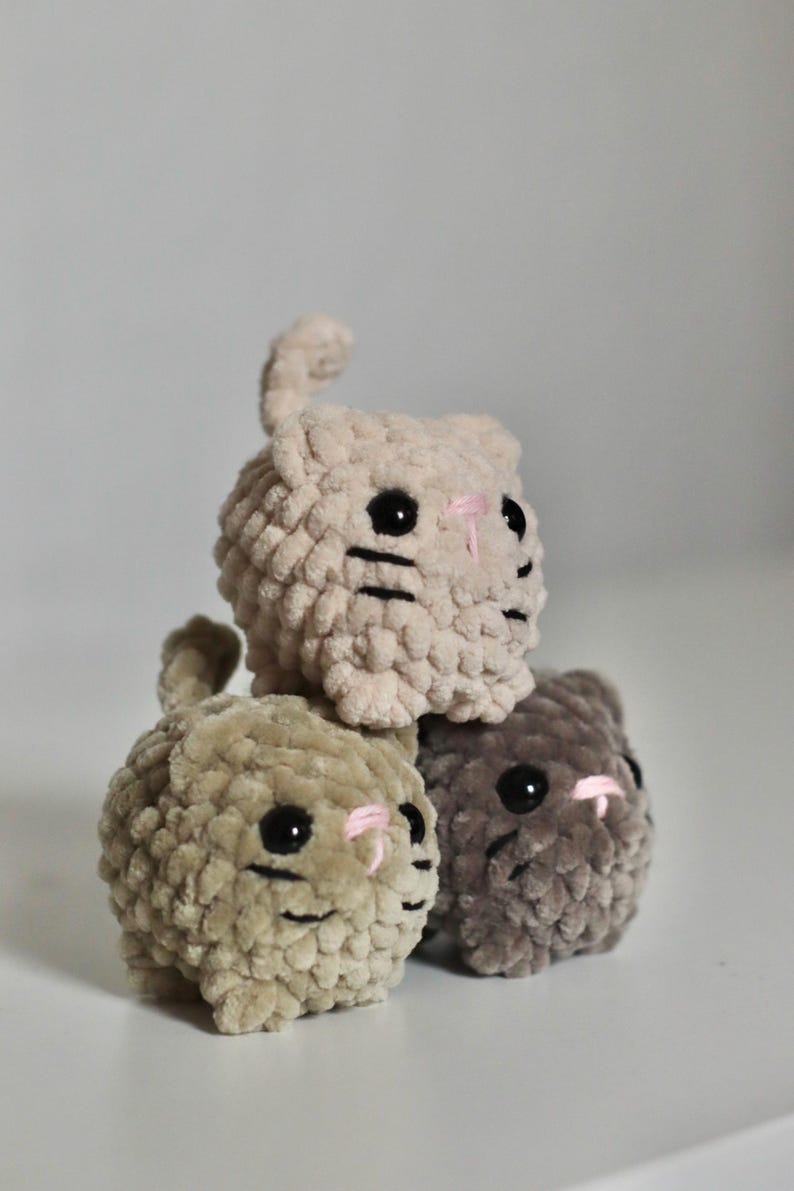 May include: Three handmade, plush cat toys in a stack. The cats are beige, olive green, and gray, with black eyes, pink noses, and black whiskers. The toys are made of a soft, textured material.
