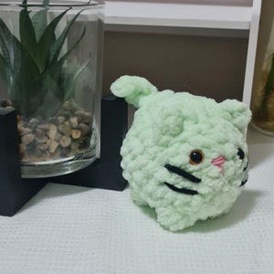 May include: A light green crocheted cat with black whiskers and a pink nose. The handmade cat has a round body and small ears. The cat is sitting on a white textured surface.