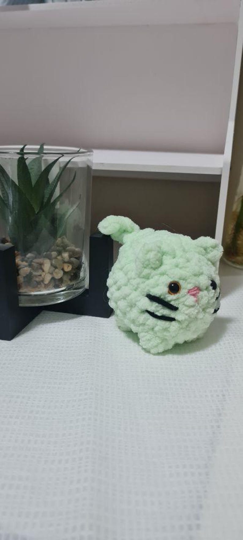 May include: A light green crocheted cat with black whiskers and a pink nose. The handmade cat has a round body and small ears. The cat is sitting on a white textured surface.