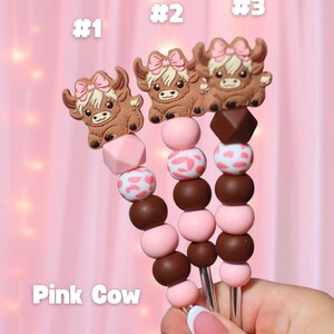 May include: Three decorative skewers with brown cow toppers wearing pink bows. The skewers have pink and brown beads, with some beads featuring a white and pink cow print. The text "Pink Cow" is visible.
