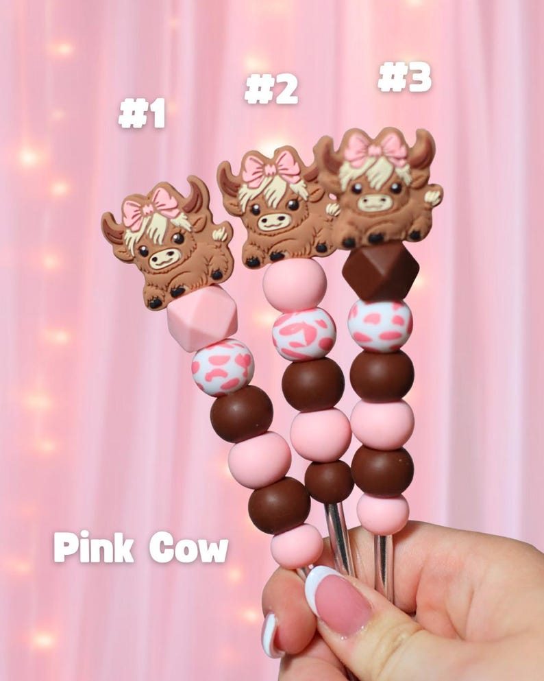 May include: Three decorative skewers with brown cow toppers wearing pink bows. The skewers have pink and brown beads, with some beads featuring a white and pink cow print. The text "Pink Cow" is visible.