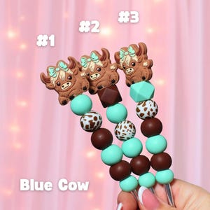 May include: Three beaded pacifier clips with brown cow-shaped charms and the text "Blue Cow". The beads are brown, white, and turquoise. Each clip is labeled with a number: #1, #2, and #3.