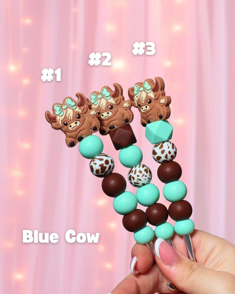 May include: Three beaded pacifier clips with brown cow-shaped charms and the text "Blue Cow". The beads are brown, white, and turquoise. Each clip is labeled with a number: #1, #2, and #3.