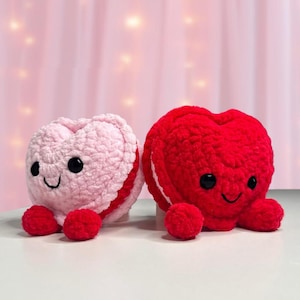 May include: Two crocheted heart-shaped plush toys. One is light pink with red accents and the other is red. Both have black button eyes and smiling faces. The toys are on a white surface with a pink background.