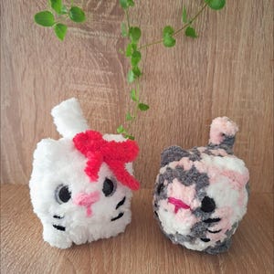 May include: Two handmade crochet cat toys. One is white with a red bow, pink nose, and black eyes and whiskers. The other is a mix of gray, pink, and white yarn with a pink nose and black eyes and whiskers. The toys are on a wooden surface.