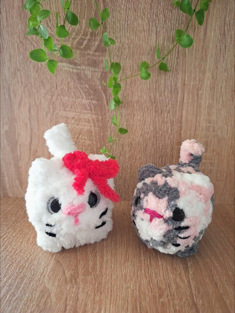 May include: Two handmade crochet cat toys. One is white with a red bow, pink nose, and black eyes and whiskers. The other is a mix of gray, pink, and white yarn with a pink nose and black eyes and whiskers. The toys are on a wooden surface.