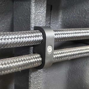 May include: A black plastic cable clamp with a screw for securing two braided cables. The clamp is attached to a gray surface.