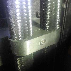 May include: Close-up of a black braided cable secured with a silver metal clamp. The clamp is attached to a black surface with a screw.