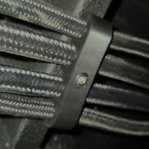 May include: Close-up of a black cable tie securing multiple black braided hoses. The tie is made of a dark material and is fastened with a single screw. The hoses are tightly bundled together.