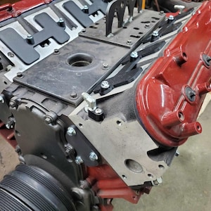 May include: Close-up of a red engine block with a black valve cover and a silver metal bracket. The engine is partially disassembled, showing the internal components.