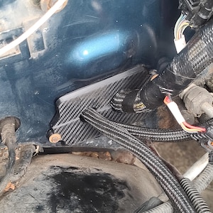 May include: A carbon fiber component installed in an engine bay. The component is black with a woven pattern and is secured with a copper-colored fastener. Various wires and hoses are also visible, along with the dark blue engine bay.
