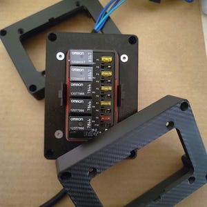May include: A black electrical component with multiple fuses and wiring, featuring the "OMRON" and "EATON" brand names. The component is mounted within a black frame, with additional black frames visible. Blue and green wires are also present.