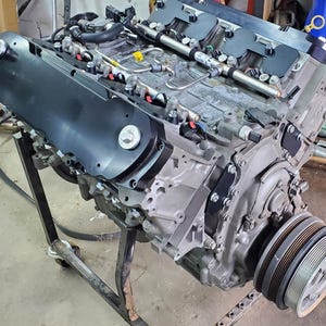 May include: A detailed view of a vehicle engine, showcasing its intricate components. The engine is primarily silver with black accents, including a prominent black cover. Various tubes and wires are visible, along with a pulley system. The engine is mounted on a black metal stand.