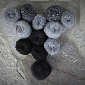 May include: An assortment of yarn balls in various shades of gray and black, arranged on a stone surface. The yarn appears soft and fluffy, with some balls having a sparkly, metallic thread woven in. The arrangement suggests a craft or knitting project.