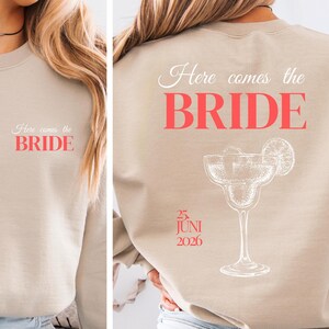 Könnte beinhalten: Beiges Sweatshirt mit dem Text "Here comes the BRIDE" in Weiß und Koralle. Auf der Rückseite befindet sich derselbe Text, eine Margarita-Illustration und das Datum "25. JUNI 2026". Ein Kleidungsstück zum Thema Hochzeit.