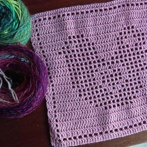 Könnte beinhalten: Ein rosa gehäkeltes Quadrat mit einem zarten Muster. Im Hintergrund sind zwei Wollknäuel zu sehen, eines grün und eines lila.