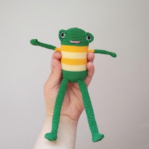 Puede incluir: Una muñeca de rana verde hecha a mano con un torso a rayas amarillas y blancas, brazos extendidos y piernas largas. La rana tiene ojos negros y boca rosa. La rana es un juguete artesanal y caprichoso.