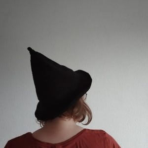 May include: A black witch hat with a pointed crown and a wide brim. The hat is made of a dark material and is worn by a person with auburn hair and a red top. The hat is a classic Halloween costume accessory.
