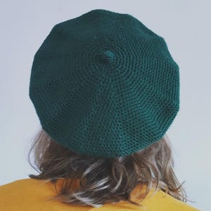 May include: A dark green beret made of a knitted material. The beret is round with a small, raised detail at the top. The beret is worn by a person with blonde hair and a yellow shirt.