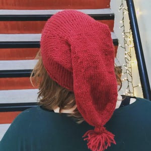 Peut inclure: Un bonnet rouge tricoté avec une longue pointe et un pompon. Le chapeau est fait d'un fil épais et texturé et semble fait à la main. Le chapeau est porté par une personne, avec le dos de sa tête et ses épaules visibles.