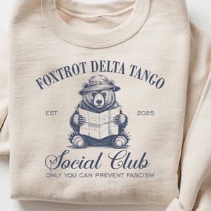 Puede incluir: Una sudadera beige con un gr&aacute;fico azul de un oso con un sombrero y sosteniendo un mapa. El gr&aacute;fico incluye el texto "Foxtrot Delta Tango Social Club Est 2023" y "Only You Can Prevent Fascism".