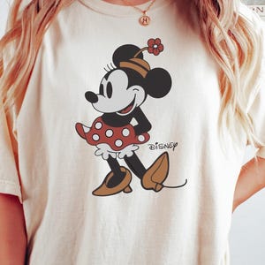 May include: Cream-colored t-shirt featuring a cartoon Minnie Mouse design. Minnie is depicted in a red polka-dot skirt, brown shoes, and a matching hat. The word "Disney" is printed below the character. The word "NATUREL" is printed at the bottom.