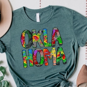 May include: Teal blue t-shirt with a colorful graphic that says "OKLA HOMA". The shirt is laid out on a white surface with a hat, jeans, and shoes.