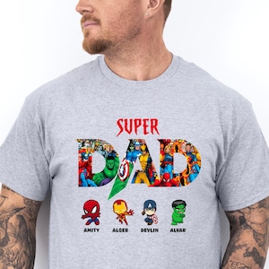 May include: A light grey t-shirt featuring the text "SUPER DAD" in red, with a collage of superhero characters forming the letters. Below, four cartoon superhero figures are labelled Amity, Alger, Devlin, and Alvar. The shirt is made of a soft material.