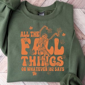 May include: An olive green sweatshirt featuring the orange text "ALL THE FALL THINGS OR WHATEVER 182 SAYS" with a skeleton and leaf graphic. The garment is folded on a light wood-effect surface.