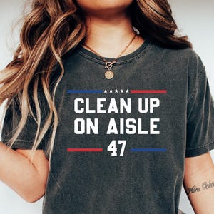 May include: A dark grey t-shirt with the text "CLEAN UP ON AISLE 47" printed in white. The text is framed by red and blue lines.