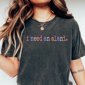 May include: A woman wearing a dark gray t-shirt with the text "i need an alani." in rainbow colors. She is wearing blue jeans and white sneakers.
