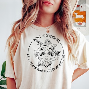 May include: Cream-colored t-shirt featuring a black and white illustration of Medusa with snakes for hair. The text around the image reads, "I WON'T BE REMEMBERED AS A WOMAN WHO KEPT HER MOUTH SHUT."