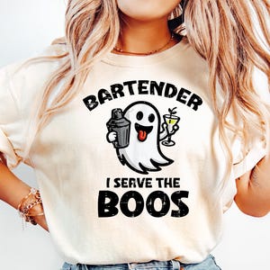 Pode incluir: Camiseta creme com o texto preto "BARTENDER I SERVE THE BOOS". Um desenho de um fantasma segurando uma coqueteleira e um copo de martini está no centro. A camiseta é combinada com jeans azul claro.
