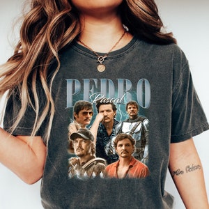 May include: A dark gray t-shirt with a vintage-style graphic featuring the text "PEDRO Pascal" and images of the actor in different roles.