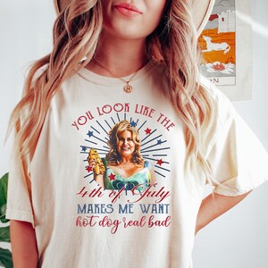 May include: A white t-shirt with a graphic print of a woman holding a hot dog. The text on the shirt reads "You look like the 4th of July makes me want hot dog real bad".