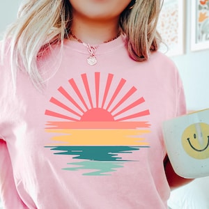 May include: A pink t-shirt with a retro sunset graphic featuring a red sun, yellow, orange, and blue stripes, and a teal and green reflection in the water.