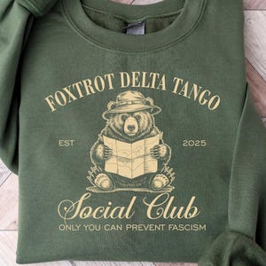 Puede incluir: Sudadera verde con un gr&aacute;fico de un oso con un sombrero y sosteniendo un mapa. El texto "Fox Trot Delta Tango Social Club Est 2025 Only You Can Prevent Fascism" est&aacute; impreso en la sudadera.