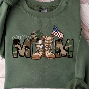 May include: A green sweatshirt with the text "army mom" in camouflage print. The design includes a pair of camouflage boots, a military hat, and a small American flag.