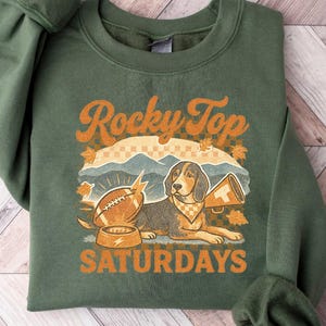 May include: Green sweatshirt with the text "Rocky Top Saturdays" in orange. The graphic features a dog, football, and megaphone. The sweatshirt is folded on a wooden surface.