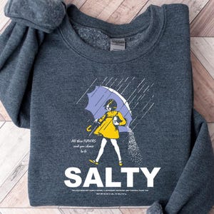 May include: Dark gray sweatshirt with a graphic of a person under a purple umbrella, wearing a yellow dress, and pouring salt. The word "SALTY" is in large white letters below the illustration. Text above the illustration reads "All the flavors and you choose to be."
